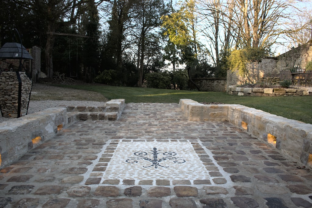 Image montrant une magnifique allée de prestige en pavés, entourés de murs en pierres, intégrant des luminaires qui sublime une mosaîque en pierre.